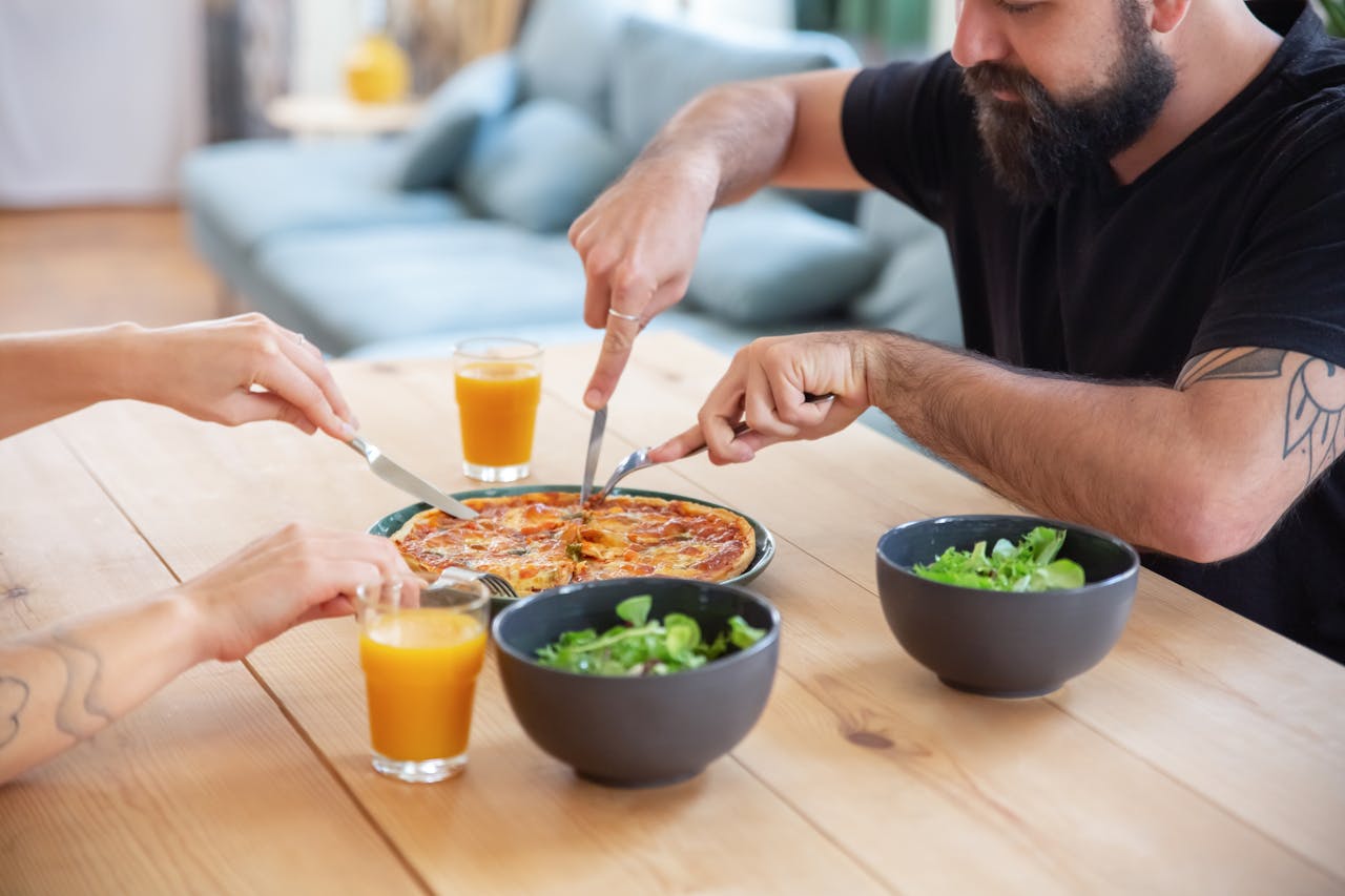 Three people enjoying pizza with orange juice and salad at a wooden table indoors.